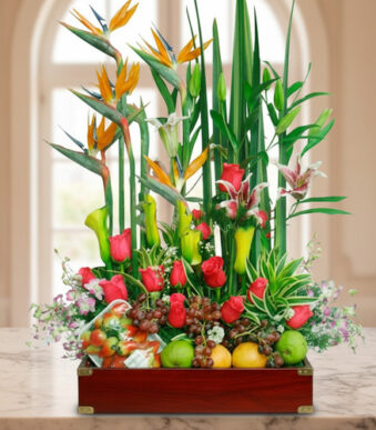 Arreglo de flores tropicales y rosas rojas combinado con frutas variadas en una caja de madera sobre una mesa de mármol, con fondo interior desenfocado