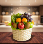 Cesta tejida de mimbre llena de frutas variadas como uvas verdes y moradas, manzanas rojas, naranjas, kiwi y peras sobre una mesa de madera con una cocina desenfocada de fondo