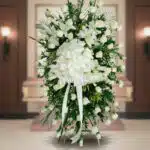 Arreglo fúnebre vertical de rosas blancas con lazo grande blanco frente a un ataúd en una sala velatoria