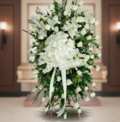 Arreglo fúnebre vertical de rosas blancas con lazo grande blanco frente a un ataúd en una sala velatoria