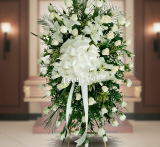 Arreglo fúnebre vertical de rosas blancas con lazo grande blanco frente a un ataúd en una sala velatoria