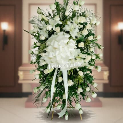 Arreglo fúnebre vertical de rosas blancas con lazo grande blanco frente a un ataúd en una sala velatoria