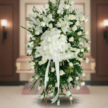 Arreglo fúnebre vertical de rosas blancas con lazo grande blanco frente a un ataúd en una sala velatoria