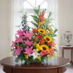 Arreglo de flores variadas con girasoles, lirios rosados y aves del paraíso sobre una mesa redonda de madera en una sala luminosa