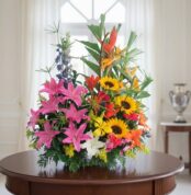 Arreglo de flores variadas con girasoles, lirios rosados y aves del paraíso sobre una mesa redonda de madera en una sala luminosa