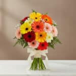 Ramo redondo de gerberas de colores rojo, naranja, amarillo, rosa y blanco con follaje verde y lazo crema, colocado sobre una mesa de mármol blanco con fondo beige