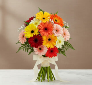 Ramo redondo de gerberas de colores rojo, naranja, amarillo, rosa y blanco con follaje verde y lazo crema, colocado sobre una mesa de mármol blanco con fondo beige
