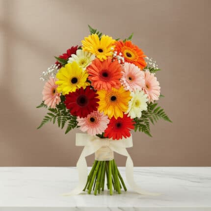 Ramo redondo de gerberas de colores rojo, naranja, amarillo, rosa y blanco con follaje verde y lazo crema, colocado sobre una mesa de mármol blanco con fondo beige
