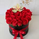Arreglo floral de rosas rojas en caja redonda negra con lazo rojo y una orquídea blanca en la parte superior