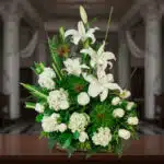 Arreglo floral de lirios blancos, rosas blancas y hortensias con follaje verde intenso, colocado sobre una mesa de madera en interior elegante con columnas al fondo desenfocado