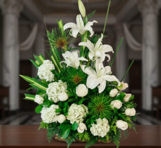 Arreglo floral de lirios blancos, rosas blancas y hortensias con follaje verde intenso, colocado sobre una mesa de madera en interior elegante con columnas al fondo desenfocado