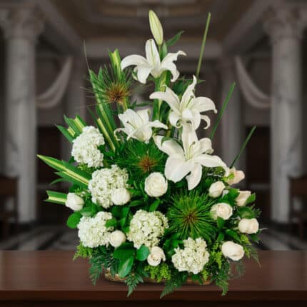 Arreglo floral de lirios blancos, rosas blancas y hortensias con follaje verde intenso, colocado sobre una mesa de madera en interior elegante con columnas al fondo desenfocado