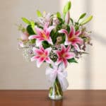 Arreglo de lirios rosas y flores blancas en un florero de cristal con lazo sobre mesa de madera