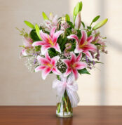 Arreglo de lirios rosas y flores blancas en un florero de cristal con lazo sobre mesa de madera