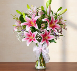 Arreglo de lirios rosas y flores blancas en un florero de cristal con lazo sobre mesa de madera