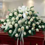 Arreglo grande de rosas blancas con lazos y follaje verde colocado sobre un ataúd de madera barnizada en una sala interior