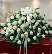 Arreglo grande de rosas blancas con lazos y follaje verde colocado sobre un ataúd de madera barnizada en una sala interior