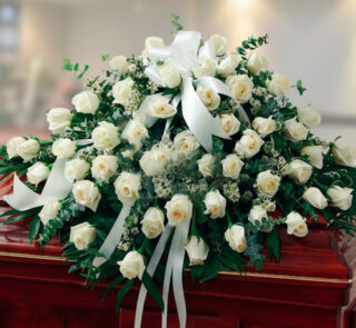 Arreglo grande de rosas blancas con lazos y follaje verde colocado sobre un ataúd de madera barnizada en una sala interior