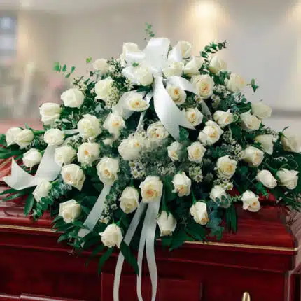 Arreglo grande de rosas blancas con lazos y follaje verde colocado sobre un ataúd de madera barnizada en una sala interior