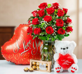 Ramo de rosas rojas en jarrón de cristal junto a globo rojo en forma de corazón con texto I Love You, oso de peluche blanco con cojín rojo y caja de chocolates Ferrero Rocher sobre mesa clara
