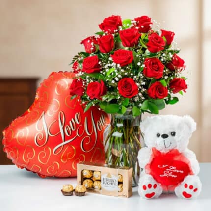 Ramo de rosas rojas en jarrón de cristal junto a globo rojo en forma de corazón con texto I Love You, oso de peluche blanco con cojín rojo y caja de chocolates Ferrero Rocher sobre mesa clara