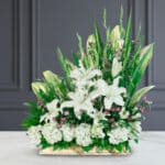 Arreglo floral rectangular con lirios blancos, flores pequeñas rosas y abundante follaje verde sobre mesa de mármol blanca