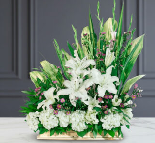 Arreglo floral rectangular con lirios blancos, flores pequeñas rosas y abundante follaje verde sobre mesa de mármol blanca