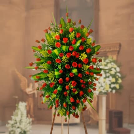 arreglo floral funerario de rosas rojas y hojas verdes sobre caballete de madera en el interior de una iglesia desenfocada