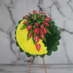 Arreglo floral circular sobre caballete de madera con media luna de flores amarillas y anthuriums rojos rodeados de hojas verdes, fondo de mármol gris