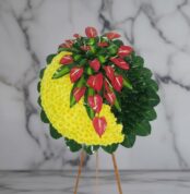 Arreglo floral circular sobre caballete de madera con media luna de flores amarillas y anthuriums rojos rodeados de hojas verdes, fondo de mármol gris