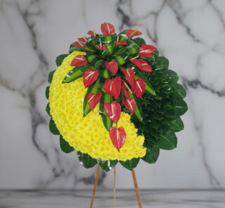 Arreglo floral circular sobre caballete de madera con media luna de flores amarillas y anthuriums rojos rodeados de hojas verdes, fondo de mármol gris