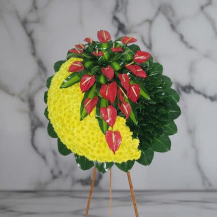 Arreglo floral circular sobre caballete de madera con media luna de flores amarillas y anthuriums rojos rodeados de hojas verdes, fondo de mármol gris