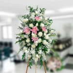 Arreglo floral funerario alto sobre caballete con lirios rosas y flores blancas en interior difuminado