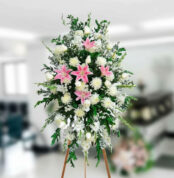 Arreglo floral funerario alto sobre caballete con lirios rosas y flores blancas en interior difuminado