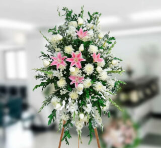 Arreglo floral funerario alto sobre caballete con lirios rosas y flores blancas en interior difuminado