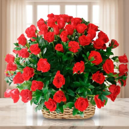 Canasta de mimbre sobre mesa de mármol con gran ramo de rosas rojas y follaje verde frente a ventana