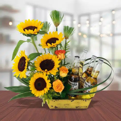Arreglo floral con girasoles y cervezas sobre mesa de madera Centro de mesa con girasoles amarillos, rosas naranjas y cuatro botellas de cerveza en base rectangular amarilla sobre mesa de madera en interior desenfocado