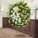 Arreglo floral redondo de pie con rosas amarillas, flores blancas y follaje verde sobre caballete de madera en interior elegante