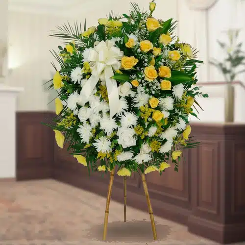 Corona floral de pie con rosas amarillas y flores blancas Arreglo floral redondo de pie con rosas amarillas, flores blancas y follaje verde sobre caballete de madera en interior elegante