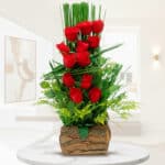 arreglo floral vertical con rosas rojas y follaje verde en base de corteza de madera sobre mesa blanca