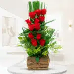 arreglo floral vertical con rosas rojas y follaje verde en base de corteza de madera sobre mesa blanca