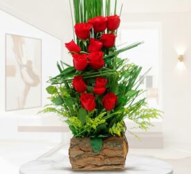 arreglo floral vertical con rosas rojas y follaje verde en base de corteza de madera sobre mesa blanca