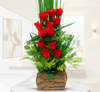 arreglo floral vertical con rosas rojas y follaje verde en base de corteza de madera sobre mesa blanca