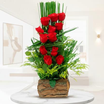 arreglo floral vertical con rosas rojas y follaje verde en base de corteza de madera sobre mesa blanca