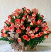canasta rectangular de mimbre con arreglo floral de rosas bicolor naranja y crema y orquídeas rosadas con follaje verde y lazo decorativo marrón