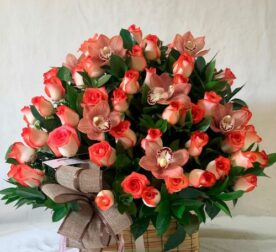 canasta rectangular de mimbre con arreglo floral de rosas bicolor naranja y crema y orquídeas rosadas con follaje verde y lazo decorativo marrón