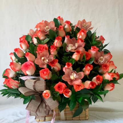 canasta rectangular de mimbre con arreglo floral de rosas bicolor naranja y crema y orquídeas rosadas con follaje verde y lazo decorativo marrón