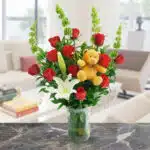 Ramo de rosas rojas y lirios blancos en florero de vidrio verde con oso de peluche amarillo, sobre mesa de mármol en sala moderna