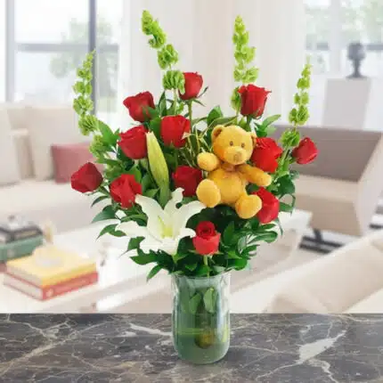 Ramo de rosas rojas y lirios blancos en florero de vidrio verde con oso de peluche amarillo, sobre mesa de mármol en sala moderna