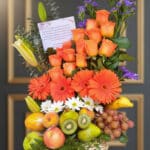 Arreglo floral con rosas y gerberas naranjas, margaritas blancas y flores moradas acompañado de canasta de frutas con manzanas, peras, kiwi, uvas, plátano y otras frutas frescas